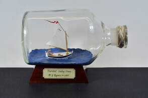 Model in a bottle of a nutshell sailing pram with a little boy in a striped shirt with a straw hat steering the tiny sailboat.  It is displayed on a wooden support with a label indicating the artist Gabrielle Rogers.